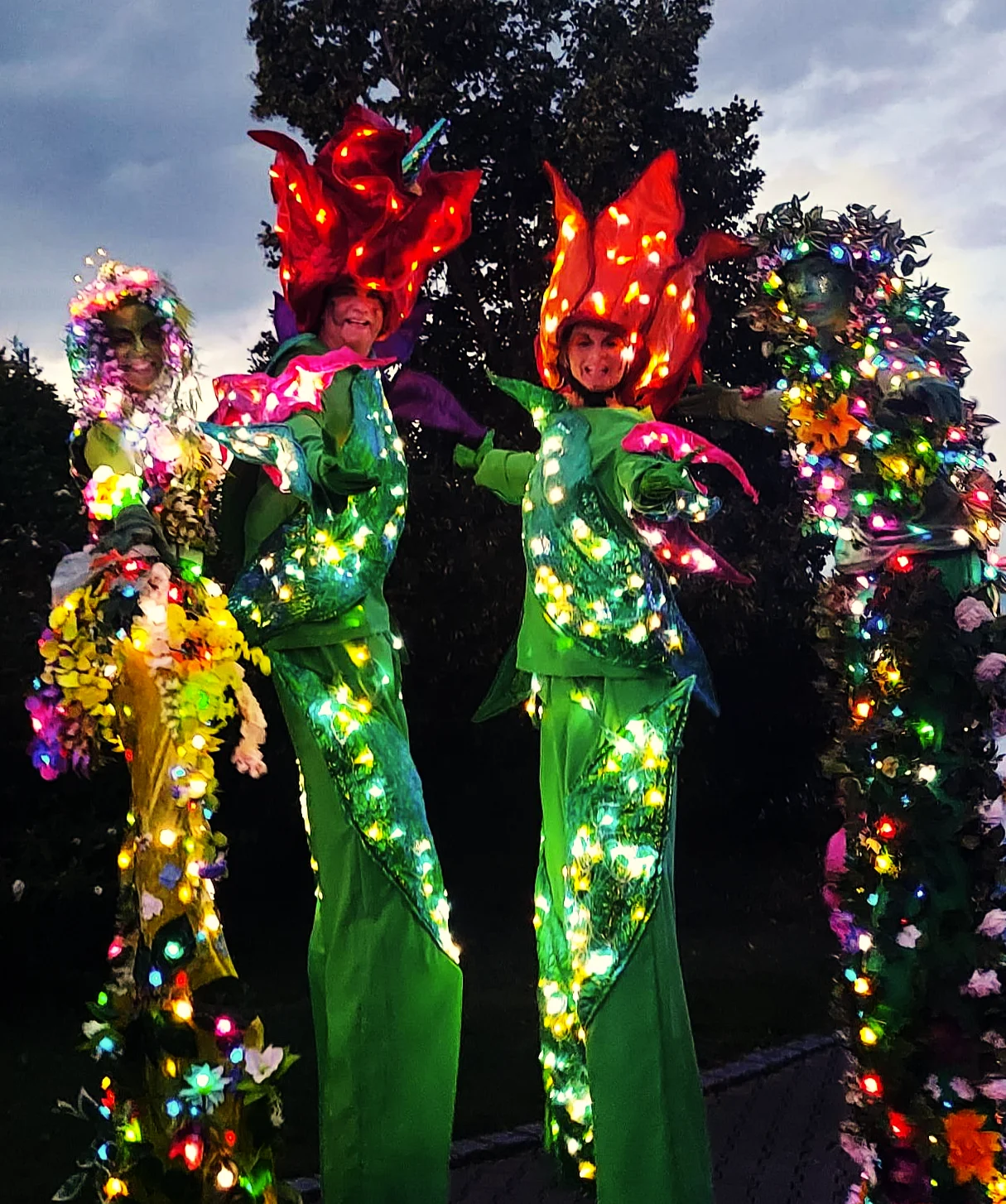 Vier Blumen auf Stelzen, Stelzengeher-Artisten von Williams Entertainment in bunten, blumigen Kostümen mit großen Blüten-Hüten und LED-Akzenten